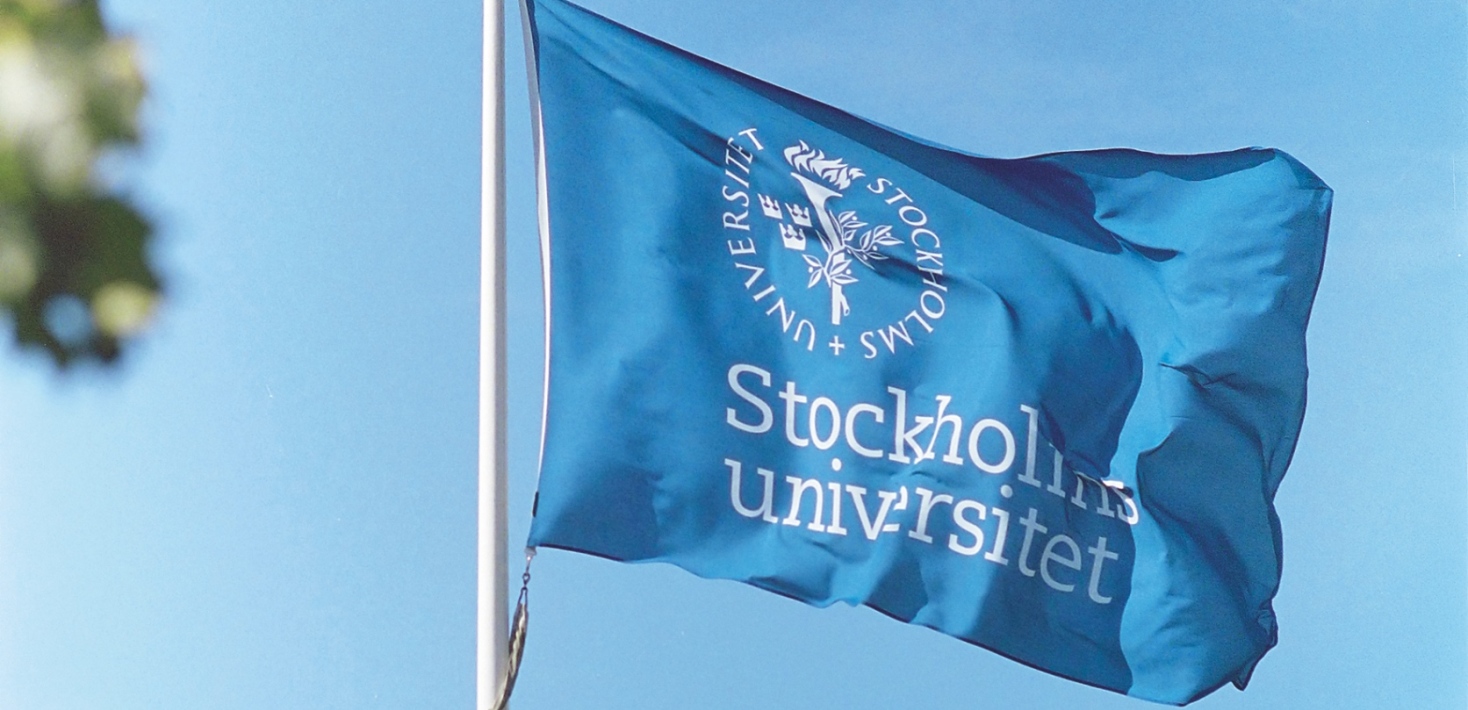 En blå flagga med Stockholms universitets vita logotyp vajar i vinden. Foto: Emanuel Almborg